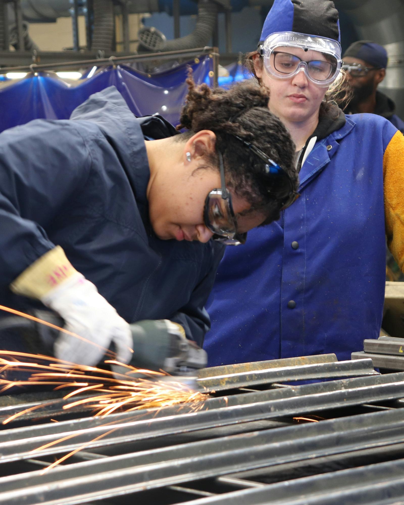 A young apprentice grinds metal while an instructor in welding gear observes